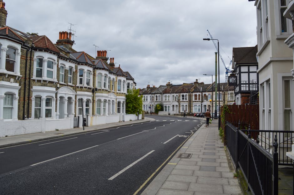 A quiet residential street with a row of Victorian-style terraced houses featuring white exteriors, bay windows, and brick chimneys under an overcast sky. The pavement runs along the right side, lined
