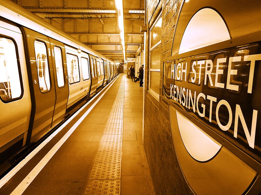 Interior view of a London Underground station platform at High Street Kensington, with a yellow and white train stationary on the left side. The platform has a tactile paving strip along the edge for