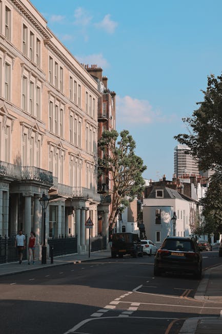 View of a residential street in Kennington with a row of Victorian-style terraced houses featuring white facades, bay windows, and red-tiled roofs. The pavement is lined with a black metal fence on on