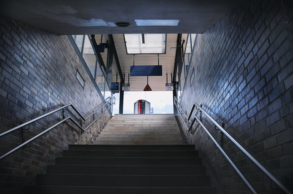 Interior view of a staircase within a building, leading downward to an underground level. The stairs are made of dark concrete with a textured surface, and each step has a raised edge. The stairway is
