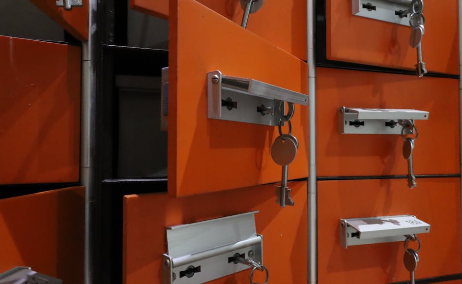 A close-up view of a set of beige metal lockers with small rectangular yellow labels and silver locks, each secured with a green key attached to a keyring. The lockers are arranged in a grid pattern, 